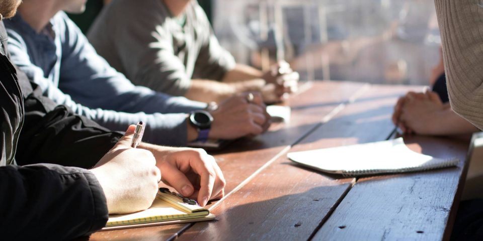 people sitting around a table in a team iinterview - the focus is on their hands and notepads in front of them, not their faces